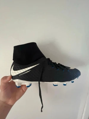 Nike Hypervenom fotbollsskor svart/vit - Säljer ett par Nike Hypervenom fotbollsskor i svart med vita detaljer och blå dobbar. Skorna har hög strumpkrage för extra stöd, snörning och en texturerad syntetisk ovandel för bättre bollkontroll. Perfekta för dig som vill ta spelet till nästa nivå. OBS!! Det är hål i strumpan 