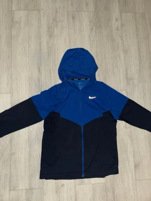 Nike miller windbreaker vindjacka - Snygg blå och svart vindjacka från Nike med huva och dragkedja framtill. Helt ny och oanvänd.