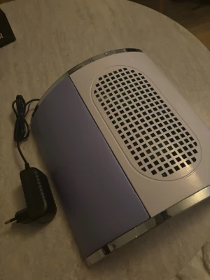 Ny Nageldammsugare med lila panel - Elektrisk nageldammsugare med vit och lila design samt silvriga detaljer. Utrustad med galler för effektiv uppsamling av nageldamm och medföljande strömadapter. Perfekt för dig som vill hålla nagelstationen ren och fräsch under manikyr.  Aldrig använt den.