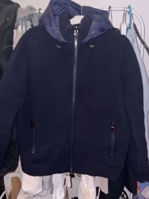 Mörkblå stickad jacka från Moncler - Säljer en mörkblå stickad jacka från Moncler med huva och dragkedja framtill. Jackan har ribbad struktur, två fickor med dragkedja och Moncler-logga på ärmen. Perfekt för kyliga dagar och riktigt snygg till vardagslooken. Storlek L men passar som M