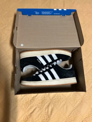 Adidas Campus svarta sneakers - Klassiska Adidas Campus sneakers i svart mocka med vita läderdetaljer och de ikoniska tre ränderna på sidan. Skorna har vit sula och vita skosnören, samt låg profil som ger en clean och tidlös look. Perfekta för dig som gillar streetwear och retrostil. Diskutera gärna pris! 