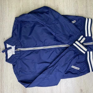 Marinblå bomberjacka från L.O.G.G. - Snygg marinblå bomberjacka från L.O.G.G. med vita och blå randiga muddar vid ärmslut och midja. Jackan har dragkedja framtill, två sidofickor med dragkedja och en klassisk bomberkrage. Materialet är lätt och passar perfekt till streetwear-stilen.