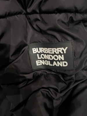 Svart pufferjacka från Burberry - Säljer en svart pufferjacka från Burberry med klassiskt rutigt innerfoder i svart, vitt och rött. Jackan har en stor huva och dragsko för justerbar passform. Perfekt för kalla vinterdagar och riktigt snygg streetstil.