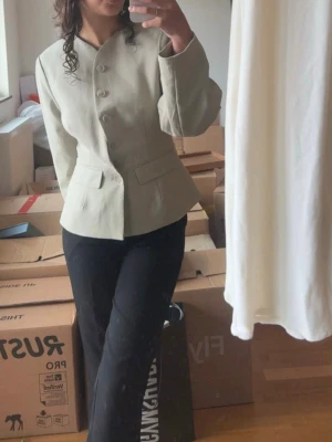 Beige kavaj med unik krage - Stilren kaki kavaj från Lounge Collection med unik asymmetrisk krage och knäppning framtill. Kavajen har två fickor med lock och markerad midja för en snygg siluett. Perfekt för dig som vill ha en modern twist på klassisk kavajstil.