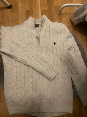 Beige kabelstickad tröja Polo Ralph Lauren - Kabelstickad beige tröja från Polo Ralph Lauren med halv dragkedja och klassisk broderad logga på bröstet. Tröjan har ribbade muddar och hög krage, perfekt för lager på lager. Materialet känns mjukt och håller värmen.