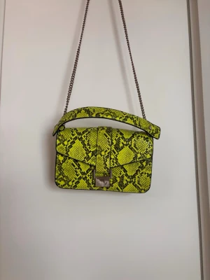 Grön väska med ormmönster - Snygg grön handväska i konstläder med ormmönster och silverfärgad kedjerem. Väskan har ett lock med metallspänne och ett praktiskt topphandtag. Perfekt statement piece för dig som vill sticka ut med din stil.
