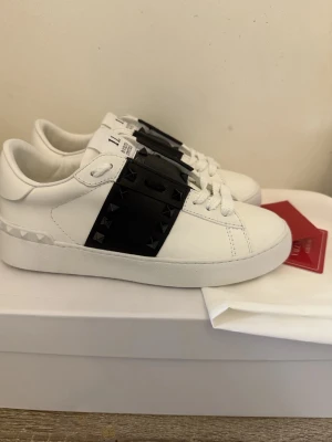 Valentino vita sneakers med nitar - Snygga vita sneakers från Valentino med svart band över vristen dekorerat med svarta nitar. Skorna är i läder och har en chunky sula samt diskreta detaljer på hälen. Perfekta för dig som vill ha en edgy och exklusiv look.