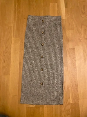 Grå ribbad långkjol med knappar - Säljer en grå ribbad långkjol med knappar framtill. Kjolen har en rak passform och är tillverkad i ett mjukt, stretchigt material som känns bekvämt. Perfekt att styla med en enkel topp eller stickad tröja.