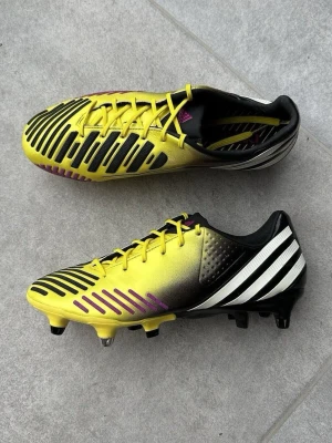 Gula Adidas Predator fotbollsskor - Snygga Adidas Predator fotbollsskor i gult och svart med vita detaljer och lila inslag. Skorna har klassisk snörning, låg profil och mönstrad ovandel för bättre bollkontroll. Yttersulan har fasta dobbar för spel på naturgräs. Perfekta för dig som vill sticka ut på planen.