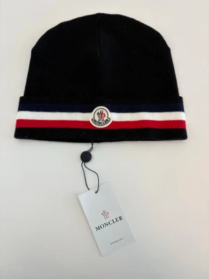 Svart Moncler mössa med ränder - Snygg svart mössa från Moncler med klassiska ränder i blått, vitt och rött runt kanten. Mössan har Moncler-logga framtill och är stickad i mjukt material. Perfekt accessoar för dig som vill ha en stilren och trendig look.