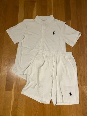 Vitt Polo Ralph Lauren set  - Säljer ett vitt Polo Ralph Lauren set med kortärmad skjorta och matchande shorts. Båda plaggen har den klassiska loggan broderad i marinblått. Skjortan har knappar framtill och shortsen har sidofickor. Perfekt för en avslappnad och stilren look.