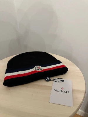 Svart Moncler mössa med ränder - Snygg svart mössa Moncler med klassiska ränder i blått, vitt och rött runt kanten. Mössan har Monclers logotyp framtill och är tillverkad i mjukt stickat material. Perfekt accessoar för att lyfta din outfit och hålla dig varm under kalla dagar.