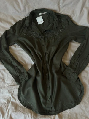 Olivgrön broderad skjorta - Snygg olivgrön skjorta med broderade detaljer vid axlar och bröst. Skjortan har lång ärm, klassisk krage och knäppning framtill. Tillverkad i ett mjukt tyg som känns skönt mot huden. Perfekt för dig som gillar stilrena men unika plagg.