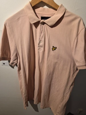 Beige/Rosa pikétröja från Lyle & Scott - Säljer en beige pikétröja från Lyle & Scott med klassisk krage och knappslå framtill. Tröjan har korta ärmar och den ikoniska gula loggan på bröstet. Perfekt för en clean och stilren look.