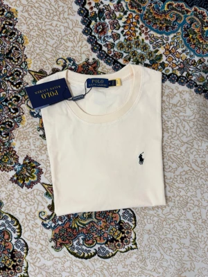 Vit t-shirt från Polo Ralph Lauren - Klassisk vit t-shirt från Polo Ralph Lauren med den ikoniska broderade loggan på bröstet. T-shirten har rund hals och är tillverkad i mjuk bomull för en skön känsla. Perfekt basplagg med stilren design.