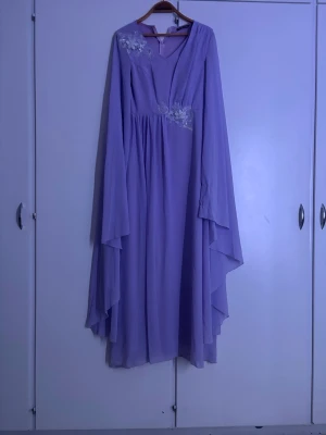 Ljuslila långklänning med spetsdetaljer - Flödande långklänning i ljuslila med v-ringning och vackra spetsapplikationer vid axel och bröst. Klänningen har vida, långa ärmar och ett mjukt, svepande fall som ger en drömmig siluett. Perfekt för dig som vill sticka ut med en romantisk look. Har aldrig använt och bara provat en gång 