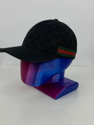 Svart keps från Gucci med GG-mönster - Snygg svart keps från Gucci med diskret GG-mönster över hela kepsen. På sidan finns en klassisk grön och röd rand. Kepsen har böjd skärm och är tillverkad i tygmaterial. Perfekt för dig som vill ha en stilren accessoar med lyxig känsla.