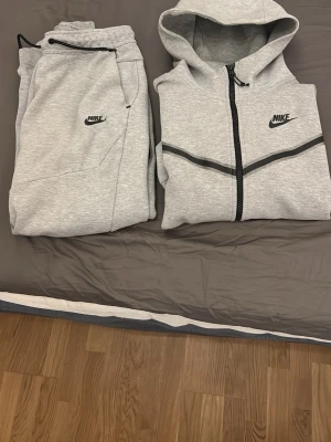 Grå zip hoodie från Nike - Snygg grå zip hoodie från Nike med svart dragkedja och svarta detaljer över bröstet. Tröjan har huva, fickor och en extra ficka på ärmen. Perfekt för en sportig och avslappnad stil. 