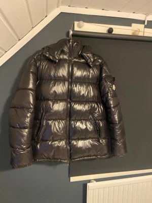 Svart glansig dunjacka från Moncler - Snygg svart dunjacka från Moncler med glansig finish och klassisk puffer-design. Jackan har hög krage, avtagbar huva och två dragkedjeförsedda fickor framtill. Moncler-logga på ärmen ger en exklusiv touch. Perfekt för kalla vinterdagar.