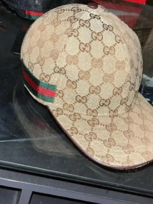 Beige keps från Gucci med GG-mönster - Säljer en beige keps från Gucci med det klassiska GG-monogrammet över hela kepsen. På sidan finns en grön och röd rand som ger en ikonisk touch. Kepsen är tillverkad i canvasmaterial och har böjd skärm. Perfekt för dig som gillar exklusiva accessoarer.