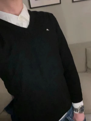 Svart stickad tröja från J.Lindeberg - Klassisk svart stickad tröja från J.Lindeberg med v-ringning och diskret logga på bröstet. Tröjan har långa ärmar och ribbade muddar vid ärmslut och nederkant. Perfekt att bära över en skjorta för en stilren look.