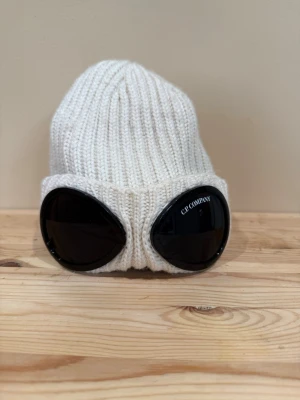 Vit stickad mössa från C.P. Company - Säljer en stilren vit stickad mössa från C.P. Company. Mössan är ribbstickad och har en klassisk form som passar perfekt till vintern. Tillverkad i mjukt material som håller dig varm och snygg under kalla dagar.