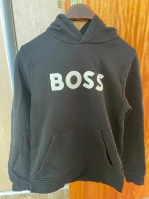 Svart hoodie med BOSS-tryck - Cool svart hoodie från BOSS med stort vitt tryck på bröstet. Hoodien har en klassisk huva och en stor magficka framtill. Perfekt för dig som gillar en enkel och stilren look. Materialet är mjukt och bekvämt, perfekt för chill dagar.Inte använd särskilt mycket inga fläckar eller så. Det står att de är en 14-S. Den passar runt 12-14 åringar,som är runt 140-150cm skulle jag säga. 