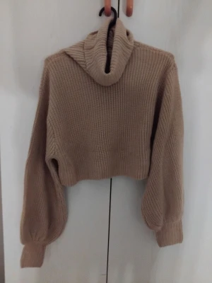 Beige croppad stickad polotröja - Trendig croppad stickad tröja i beige med hög polokrage och ballongärmar. Grovstickad i mjukt material, perfekt för lager på lager. Ribbstickade muddar vid ärmslut och nederkant ger en snygg finish.