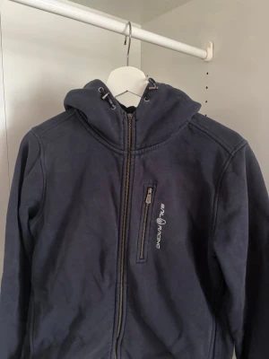 Mörkblå hoodie från Sail Racing - Säljer en mörkblå hoodie från Sail Racing med dragkedja framtill och en bröstficka med logga. Hoodien har justerbar huva med snören och är tillverkad i mjukt material, perfekt för chill dagar. Klassisk och stilren design som passar till det mesta. 