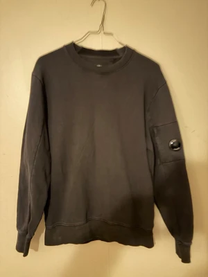 Svart sweatshirt från C.P. Company - Säljer en svart sweatshirt från C.P. Company med rund hals och lång ärm. Tröjan har en ikonisk linsdetalj på ena ärmen och ribbade muddar vid ärmslut och nederkant. Tillverkad i mjukt bomullsmaterial för en skön och avslappnad look.
