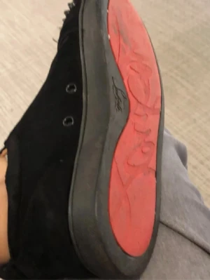 Svarta sneakers med nitar från Louboutin - Säljer ett par svarta sneakers från Christian Louboutin med klassisk röd sula och svarta nitar på tån. Skorna har snörning och är tillverkade i mocka för en lyxig känsla. Perfekta för dig som vill sticka ut med en edgy och exklusiv stil.