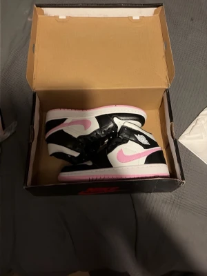 Nike Air Jordan 1 svart/vit/rosa - Säljer ett par Nike Air Jordan 1 sneakers i svart, vitt och rosa. Skorna har klassisk high-top design, rosa swoosh och yttersula, samt svart och vitt läder på ovandelen. Perfekta för dig som vill sticka ut med snygga färgdetaljer och ikonisk siluett.