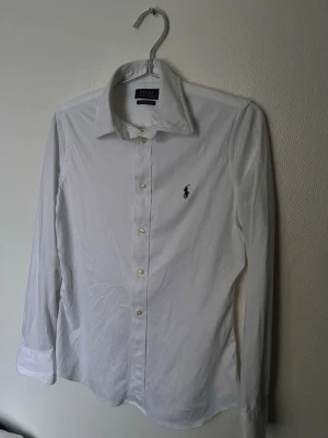 Vit skjorta från Polo Ralph Lauren - Klassisk vit skjorta från Polo Ralph Lauren med lång ärm och knappar framtill. Skjortan har den ikoniska broderade loggan i marinblått på bröstet och är tillverkad i bomull för en skön och stilren look.