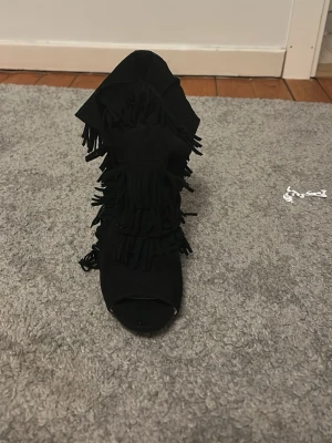 Svarta klackar med fransar - Säljer ett par svarta stövletter med hög smal klack och öppen tå. Skorna har flera lager fransar längs sidorna och framtill för en cool boho-vibe. Materialet ser ut att vara mockaimitation. Perfekta för dig som vill sticka ut!