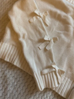 Vit stickad tröja med rosetter - Supersöt vit stickad tröja med dekorativa rosetter längs ena sidan. Tröjan har långa ärmar och ribbade muddar vid ärmslut och nederkant. Perfekt för dig som gillar gulliga detaljer och en mjuk, mysig känsla.