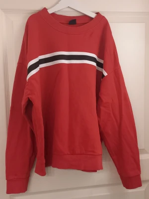 Röd sweatshirt från Gina Tricot - Röd sweatshirt från Gina Tricot med vita och svarta ränder över bröstet. Tröjan har rund halsringning, långa ärmar och en relaxed passform. Perfekt för en avslappnad och sportig stil.