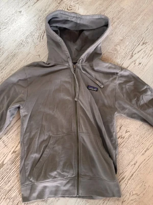 Grå zip-hoodie från Patagonia - Snygg grå hoodie från Patagonia med dragkedja framtill och klassisk huva med dragsko. Hoodien har två stora fickor fram och en liten logga på bröstet. Perfekt till jeans eller chill-outfits. Tillverkad i mjukt bomullsmaterial.