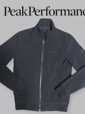 Svart vindjacka från Peak Performance - Snygg svart vindjacka från Peak Performance med hög krage och hel dragkedja framtill. Jackan har diskret logga på bröstet och ribbade muddar vid ärmslut och nederkant. Perfekt för dig som gillar stilren och sportig look.