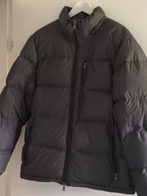 Svart pufferjacka från Polo Ralph Lauren - Säljer en svart pufferjacka från Polo Ralph Lauren med hög krage och dragkedja framtill. Jackan har en bröstficka med dragkedja och två sidofickor. Klassisk quiltad design och diskret logga på bröstet. Perfekt för kalla vinterdagar.