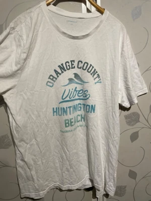 Vit t-shirt Orange County Vibes - Vit t-shirt från Airrely med tryck i blått framtill: 'Orange County Vibes Huntington Beach'. Klassisk rund hals och korta ärmar. Tillverkad i mjuk bomull, perfekt för en avslappnad och chill stil.