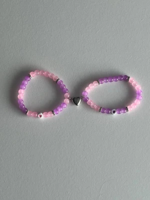 Lila och rosa pärlarmband med magnet-hjärta - Två elastiska armband med glansiga pärlor i lila och rosa, dekorerade med små silvriga detaljer. Armbanden sitter ihop med ett silverfärgat hjärthänge i mitten. Perfekt för dig som gillar söta accessoarer och vill addera färg till din stil.