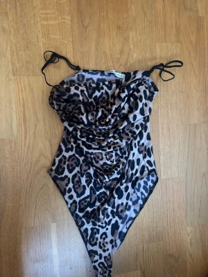 Leopardmönstrad mesh body - ALDRIG ANVÄND! Säljer en snygg body med leopardmönster och tunna svarta axelband från Zara. Materialet är mesh som ger en lätt transparent känsla och plagget har draperad front för extra stil. Perfekt för dig som vill sticka ut med ett djurmönstrat plagg.