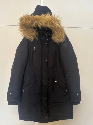 Svart jacka - Svart jacka med stor huva och beige fuskpälsdetalj. Jackan har två bröstfickor med dragkedja och två stora fickor med lock framtill. Långa ärmar med knappdetaljer och dragkedja framtill. Perfekt för kalla dagar och riktigt mysig att bära.