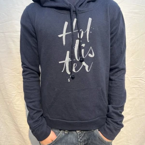 Mörkblå hoodie från Hollister - Mörkblå hoodie från Hollister med stor vit logga framtill. Klassisk känguruficka, justerbar dragsko i huvan och mjukt material. Perfekt för en avslappnad och trendig look. Storlek S men passar xs.