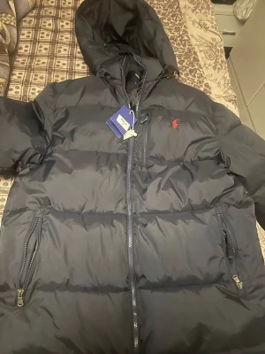 Svart pufferjacka från Polo Ralph Lauren - Snygg svart pufferjacka från Polo Ralph Lauren med huva och dragkedja framtill. Jackan har två sidofickor med dragkedja och den klassiska röda loggan broderad på bröstet. Perfekt för kalla vinterdagar och riktigt skön att bära.