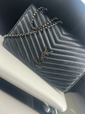 Svart  handväska med kedjerem - Säljer en svart handväska  med klassisk i silver framtill. Väskan har quiltat mönster i chevronstil och en snygg silverfärgad kedjerem. Perfekt för dig som gillar lyxiga accessoarer och tidlös design.Fake)) skriv för mer bilder 