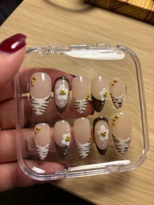 Bruna press-on naglar med blommor - Snygga press-on naglar i mandelform med beige och bruna toner, dekorerade med vita blommor och små guldfärgade pärlor. Några naglar har zebramönster och andra har fransk tipp i brunt. Perfekt för dig som vill ha en unik och trendig look. 10st naglar
