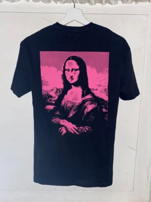 Svart t-shirt med Mona Lisa-tryck - Cool svart t-shirt från Yourturn med ett stort rosa Mona Lisa-tryck på ryggen och liten rosa logga framtill. Klassisk passform och rund hals. Perfekt för dig som gillar streetwear och grafiska motiv. Storlek XXS men i oversized så passar S, den har inte kommit till användning 