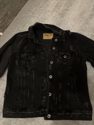 Svart jeansjacka  - Snygg svart jeansjacka i storlek S. Jackan har slitna detaljer, klassisk krage, två bröstfickor med lock och knappar samt två sidofickor. Silverfärgade knappar framtill och på ärmslut. Perfekt för en cool och avslappnad stil.
