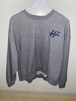 Grå sweatshirt med mörkblå logga - Säljer en grå sweatshirt med rund hals och mörkblå logga på sidan av bröstet. Tröjan har långa ärmar, ribbade muddar och en relaxed passform. Perfekt för dig som gillar enkel och stilren streetwear.